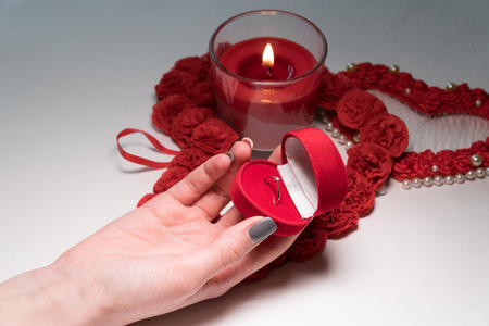 The female hand reaches for the ring in a case in the form of a heart that stands on a white table. Composition of two hearts and candles.の写真素材