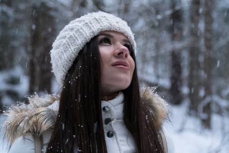 Beautiful girl with long dark hair in a white jacket walking in a winter forest during a snowfallの写真素材
