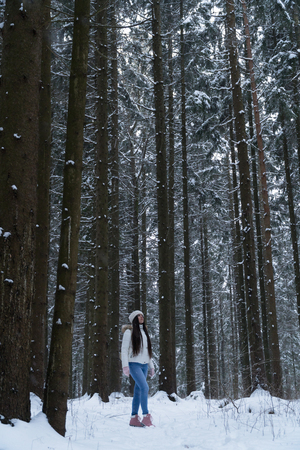 Beautiful girl with long dark hair in a white jacket walking in a winter forest during a snowfallの写真素材