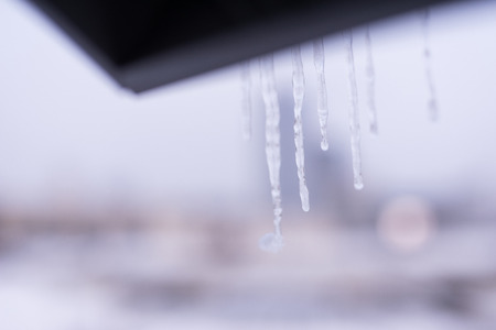 Huge icicles hang from the roof of the house. In the street there is snowfall.