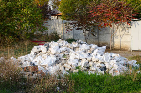 The sunlit pile of construction garbage on the green grass with uncultivated white flowers and autumn trees, fence and house on the background. Street scene.の写真素材