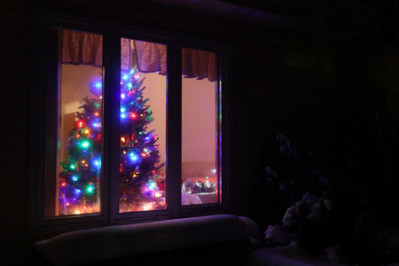Glowing Christmas tree in the window. Snowy winter night scene. Winter holidays background, Christmas background, New Year background. Good for greeting card.の写真素材