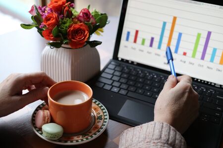 Home based remote work during new reality and quarantine, Social distancing concept. Working space view with cup of coffee with sweat treats between woman hands, working on the laptop computer and vase with beautiful flowers bouquet in a background.の写真素材