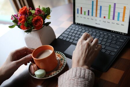 Home based remote work during new reality and quarantine, Social distancing concept. Working space view with cup of coffee with sweat treats between woman hands, working on the laptop computer and vase with beautiful flowers bouquet in a background.の写真素材