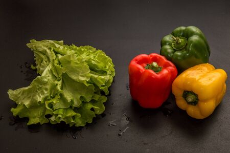 Close up, macro. Flat lay. Still life. Three multicolored peppers (paprika) and a big bunch of fresh green lettuce on black background.の写真素材