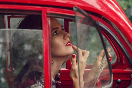 Close up. A beautiful pin-up girl in a plaid shirt corrects make-up in the salon of an old red retro car.の写真素材
