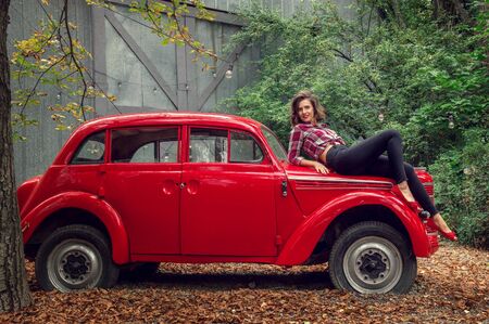 Pin-up girl in jeans and a plaid shirt is posing on a russian red retro car. She is looking at the camera and smiling happily.の写真素材