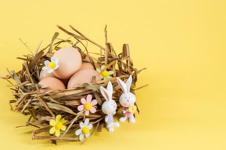 Close up, macro. Easter still life. Farm eggs in a nest of straw. Two cute porcelain bunnies in the foreground. The decor of the felt flowers. Yellow background. Copy space.の写真素材