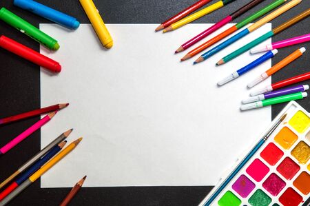 Close up, macro, flat lay. "Back to school" background. Black desk with a white unfilled sheet of paper. Felt pens, paints, pencils, crayons are scattered around. Copy space.の写真素材