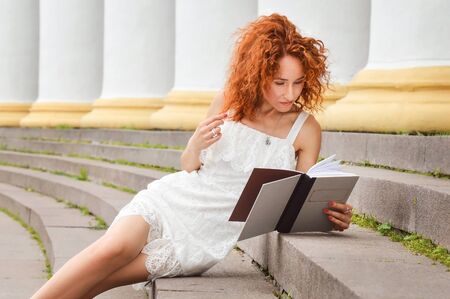 Close up. A slender red-haired girl in a white dress sits on the steps near the columns and studies. Copy space.の写真素材