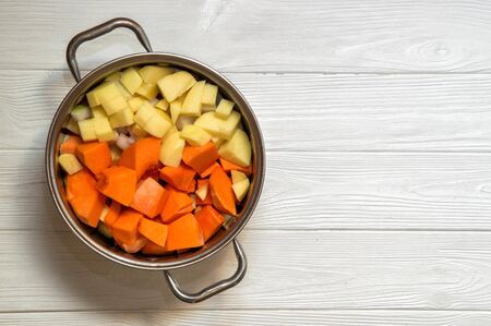 Close up, top view. Soup pot with sliced ingredients for pumpkin cream soup: carrot, pumkin, onion, potatoes. Plant-based food on the white wooden background. Copy space.の写真素材
