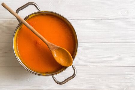 Close up, top view. Soup pot with pumpkin cream soup. A wooden spoon lies on top of the pan. Plant-based food on the white wooden background. Copy space.の写真素材