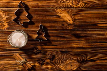 Close up, flat lay. A white candle in a glass candlestick is burning, spices and metal molds in the shape of Christmas trees are laid out around it. Rustic brown wooden background. Copy space.の写真素材