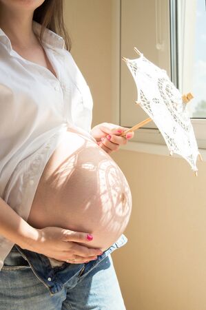 Pregnancy. A pregnant woman stands near a window and holds an openwork white umbrella. On the stomach lies a shadow in the form of a patterned flower.の写真素材