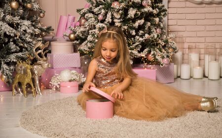 Close up. Christmas concept. A beautiful girl in an elegant golden dress sits on a soft fluffy carpet and opens a pink gift box. Christmas tree and fireplace near.の写真素材