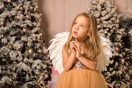 Close up. Christmas concept. A little girl in the image of a Christmas angel with wings dreamily folded her arms and looks up. She is dressed in a golden dress, standing near elegant Christmas trees.の写真素材