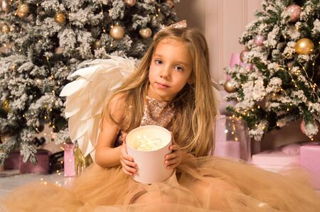 Close up. Christmas studio. A little girl in a gold dress and with angel wings holds in her hands a box with luminous lights.の写真素材