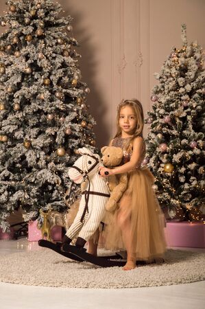 Close up. A little girl riding a toy rocking horse with a teddy bear. Festively decorated room with a Christmas tree. Christmas concept.の写真素材