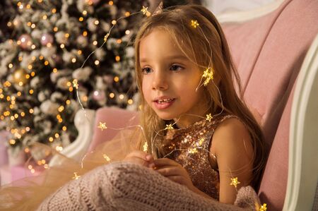 Close up. Portrait of a little beautiful girl framed by the luminous lights of a New Year's garland. Against the background of the Christmas tree. Copy space.の写真素材