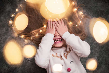 Close up. A little blonde girl is waiting for gifts, covering her eyes with her hands and laughing fervently. Gray background, Christmas garlands.の写真素材