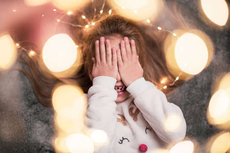Close up. A little girl is waiting for gifts, dreamily covering her eyes with her hands and laughing. Gray background, Christmas garlands.の写真素材