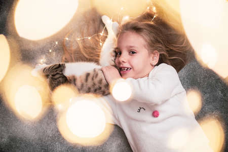 Close up. A little girl with a mischievous look is holding a kitten playing with a festive glowing garland. Gray backgroung. The frame is framed with bokeh of Christmas garland.の写真素材
