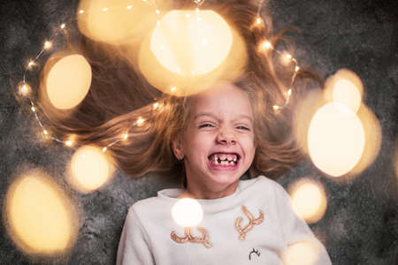 Close up. Portrait of a perky laughing 6-year-old girl framed by bokeh from a festive garland. Christmas mood.の写真素材