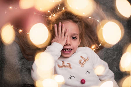 Close up. A little blonde is waiting for gifts, she closed her eyes with her palm, looks into the camera. Gray background, Christmas garlands.の写真素材