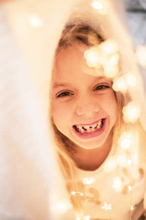 Close up. A portrait of a laughing 6-year-old girl hiding in a white plaid and festive garlands.の写真素材