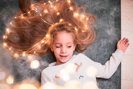 Close up. Portrait of a little girl with blond hair braided with festive garlands. She narrowed her eyes slyly and looks down for gifts.の写真素材