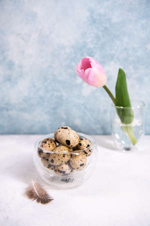 A bunch of brown quail eggs in a transparent bowl. Nearby are pink tulip. White and blue pastel background. Preparing for Easter. Copy space. Vertical oriented.の写真素材
