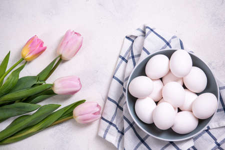 A bunch of white chicken eggs in a blue bowl. White background. Nearby are pink tulips. Preparing...