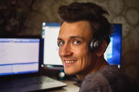 Close-up. Portrait of a smiling young man in headphones working on two computers at home.