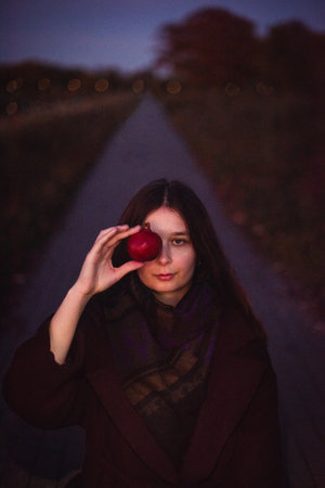 Close-up. Vertical portrait of a young girl in a burgundy coat holding a pomegranate near her face. Evening park in the background.の写真素材