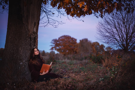 Picturesque lawn in the park. A young girl under a tree is dreaming, holding a book at sunset.の写真素材