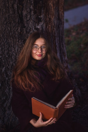 Close-up. A young girl sitting under an autumn tree and smiling dreamily. She is holding a book in her hands.の写真素材