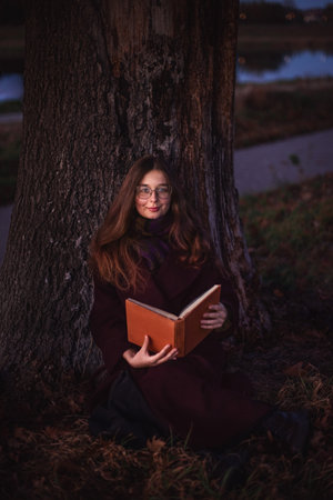 Close-up portrait. A young girl sitting under an autumn tree and smiling dreamily. She is holding a book in her hands.の写真素材