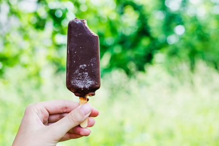 Chocolate ice cream in chocolate glaze on a stick in a female hand on a green backgroundの写真素材