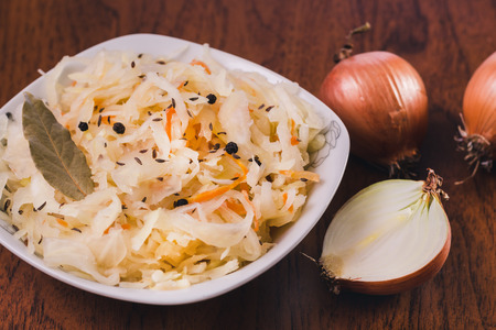 Salad of sauerkraut and carrots with laurel leaves, black pepper and cumin in a white plate and several onion bulbs on a wooden tableの写真素材