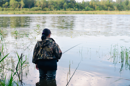 Woman with dark hair in dark clothes and high rubber boots is fishing with a fishing rod on a beautiful lakeの写真素材