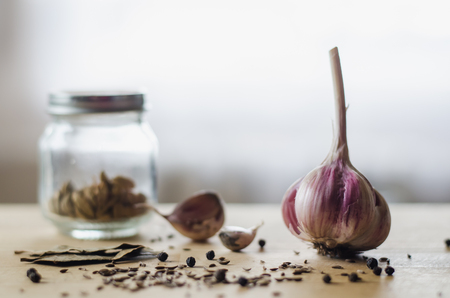 Head of garlic, black peppercorns, cumin seeds, laurel leaves and a jar of spices on a light wooden tableの写真素材