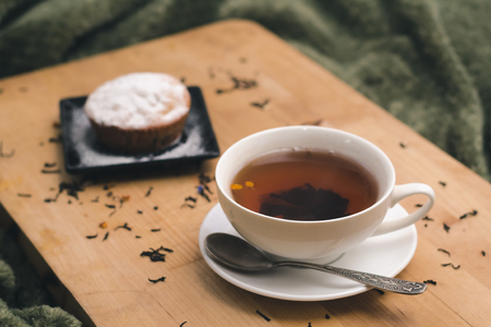 Homemade cupcake with powdered sugar on a black plate and a white cup of tea with natural additives on a wooden tray on a background of green textiles. The concept of a cozy breakfast in bedの写真素材