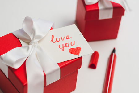 Red gift boxes tied with a white ribbon, a marker and a card with an inscription "love you" on a light background. The process of preparing a gift for a loved one.の写真素材