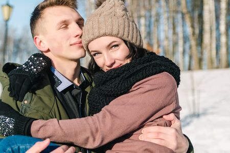 Young beautiful couple in love walking in a winter snowy park on a bright sunny day. Young man holds...