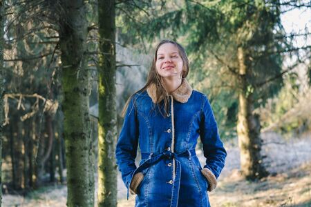 Young slim girl with long hair in a denim coat laughs in a sunny spring coniferous forestの写真素材