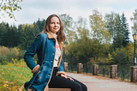 Beautiful girl with long hair in a blue denim coat sits in an autumn park