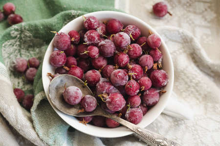 Frozen berries of red gooseberries with hoarfrost in a white cup and a spoon on an old linen tablecloth, top viewの写真素材