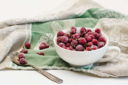 Frozen berries of red gooseberries with hoarfrost in a white cup and a spoon on an old linen tableclothの写真素材