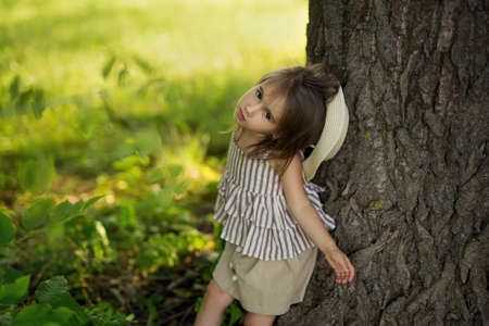 A little girl model in a hat poses for a photo at a huge tree trunk in the park.の写真素材