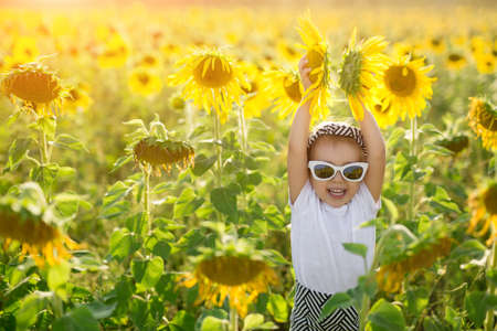 A little girl in sunglasses laughs while holding two flowers in a huge field of sunflowers at sunset.の写真素材